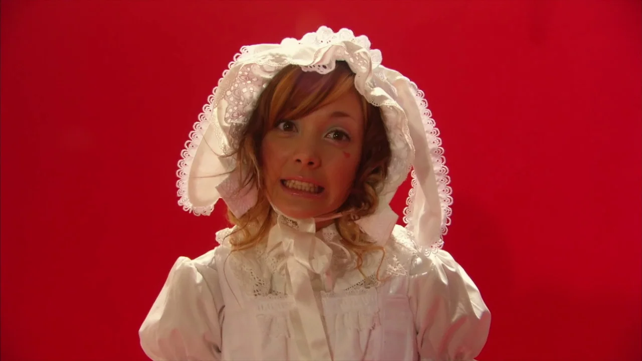 Kamikaze Girls (2004) movie still: medium — A person stands centered against a solid, brightly lit red background. The subject wears a frilly white…
