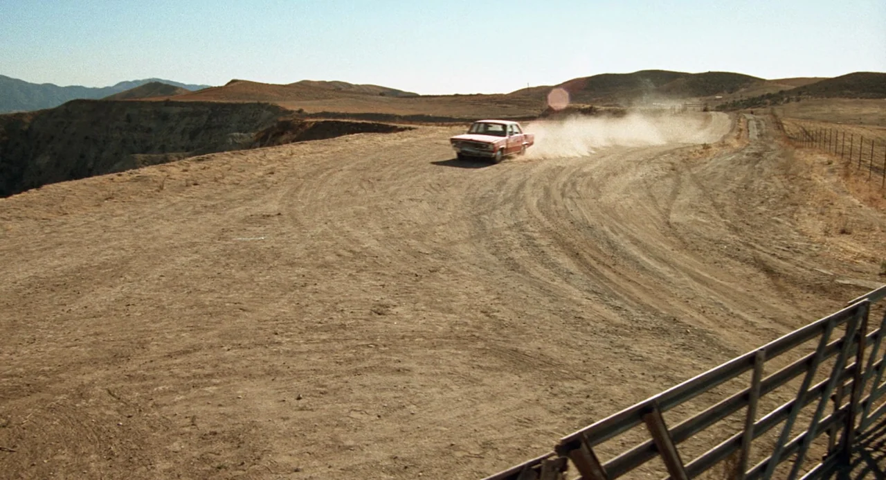 Duel (1971) movie still: wide — A dry, hilly landscape features a winding dirt road under a clear sky. A vintage red sedan travels around a bend,…