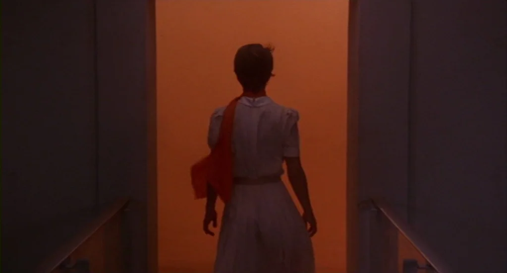 Cat People (1982) movie still: fullbody — A person walks through a dim, nondescript indoor corridor toward a glowing orange rectangular exit. The…