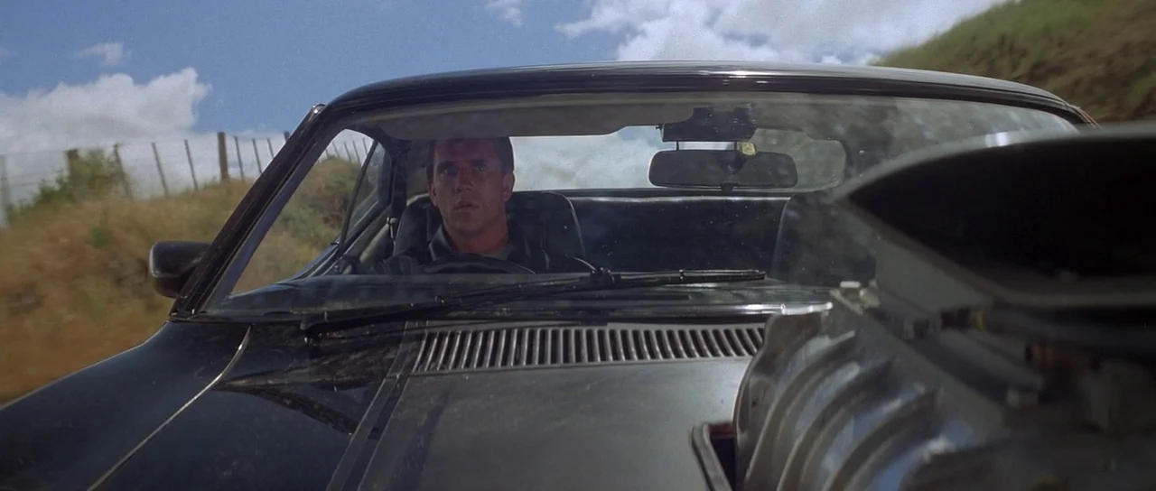 Mad Max (1979) movie still: medium — A man drives a black muscle car with a large engine blower on the hood. The car moves along a dirt road with…