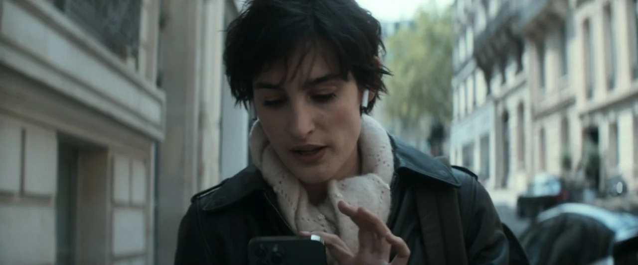I Love America (2022) movie still: medium — A Paris-style city street lined with tall apartment buildings on a bright day. A woman wearing a leather…