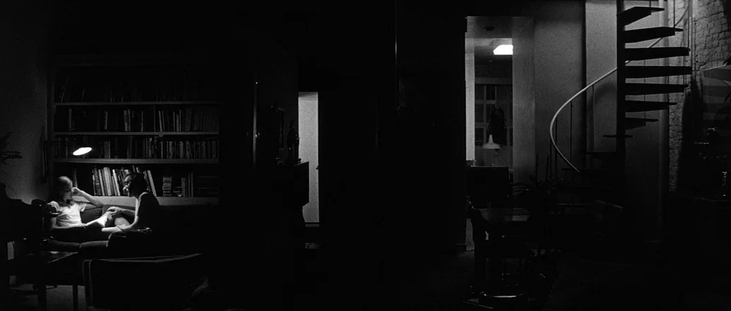Manhattan (1979) movie still: wide — A dark, spacious open-plan dwelling features a spiral staircase and a large bookshelf. Two people sit together…
