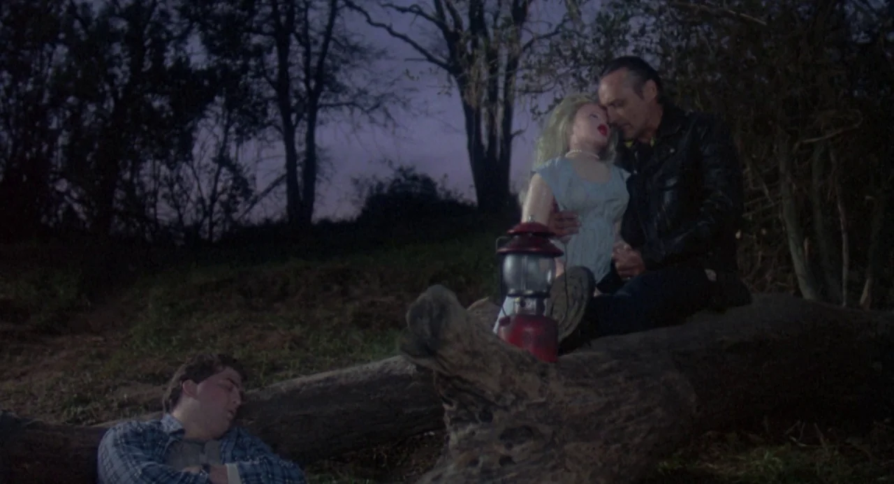River’s Edge (1986) movie still: wide — A dark wooded clearing at night with silhouetted trees against a twilight purple sky. One man sits on a…