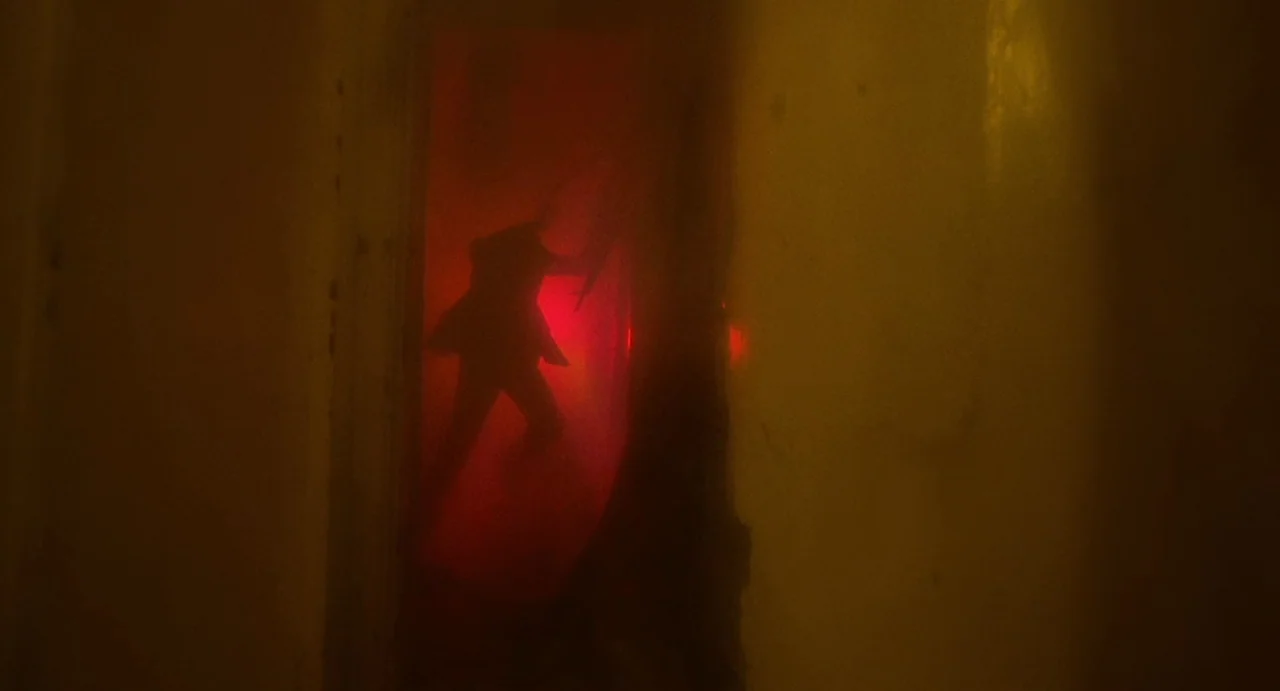 God Told Me To (1976) movie still: wide — A narrow dimly lit corridor filled with haze features a glowing red doorway. A dark silhouette of a man in…