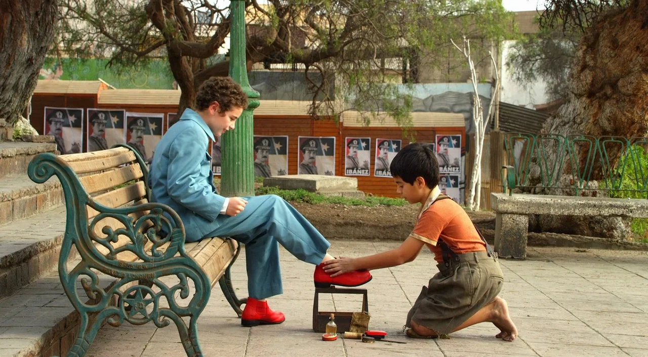 Dance of Reality (2013) movie still: medium — An outdoor park area with a paved ground surface and large, gnarled trees nearby. A boy sits on a…