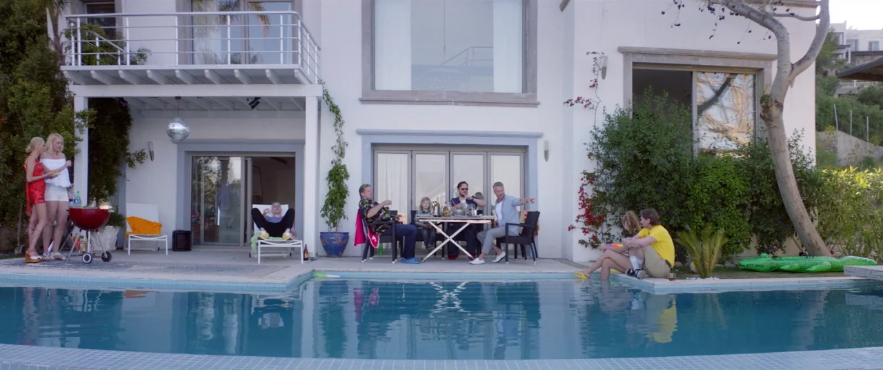 Holiday movie still: wide — A group of people socialize on the patio of a modern white villa during daylight. Some individuals recline on chairs…