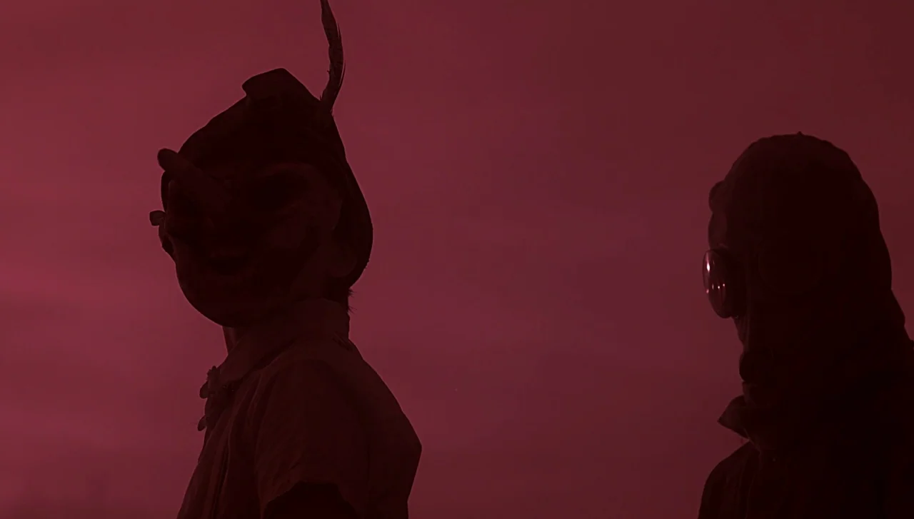 Hellions (2015) movie still: medium — Two masked individuals stand in a dimly lit, red-hued exterior environment. They are posed side-by-side,…
