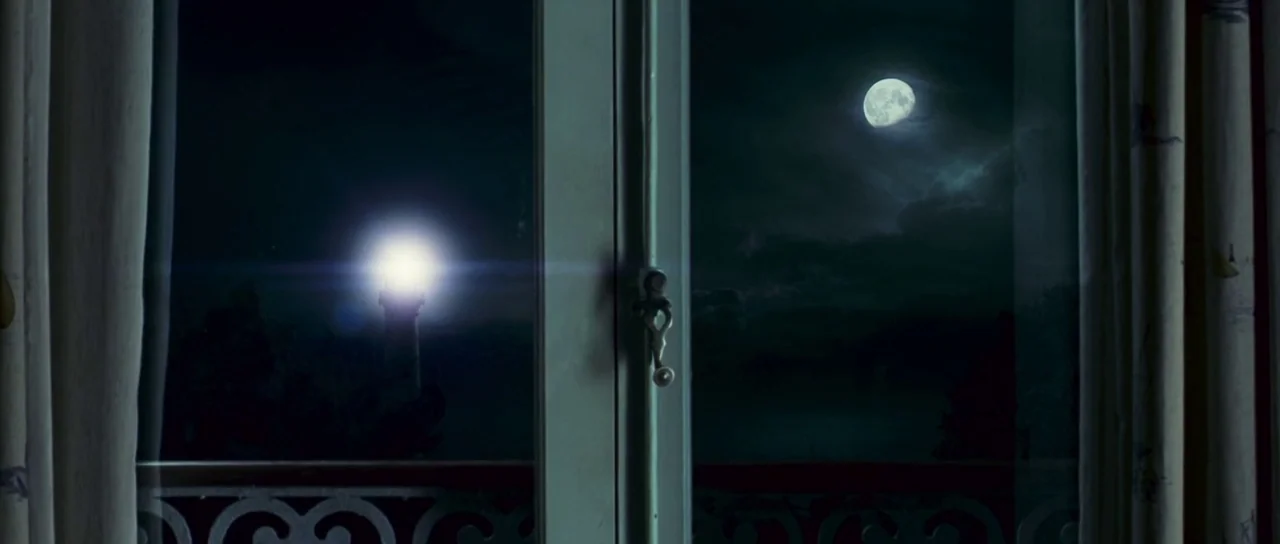 The Orphanage (2007) movie still: establishing — A view through a window frame looking out at a nocturnal landscape. A lighthouse beacon shines in…