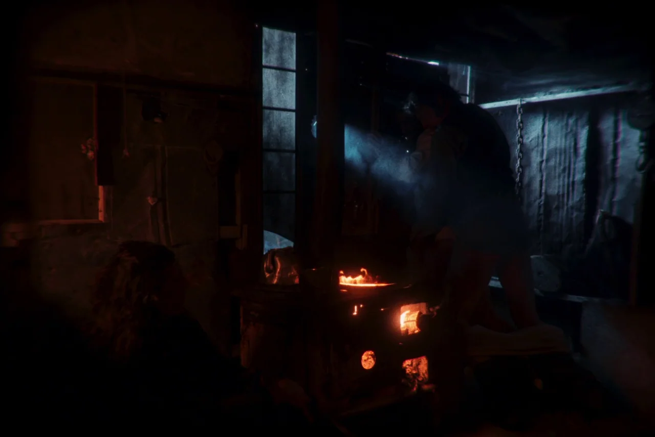 The Settlers (2023) movie still: medium — A dark room contains a cast-iron stove with visible glowing embers and orange flames. A man stands at the…