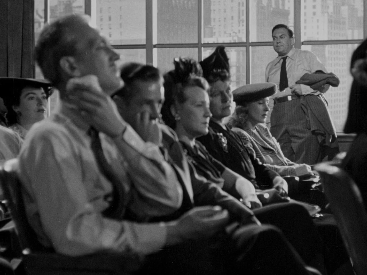 Phantom Lady (1944) movie still: medium — A group of people sit in rows in an auditorium with large windows overlooking a city. A man stands in the…