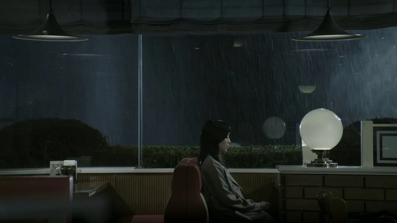 Confessions (2010) movie still: medium — A woman sits in a dark diner booth at night during a heavy downpour. Rain streaks the large glass window…
