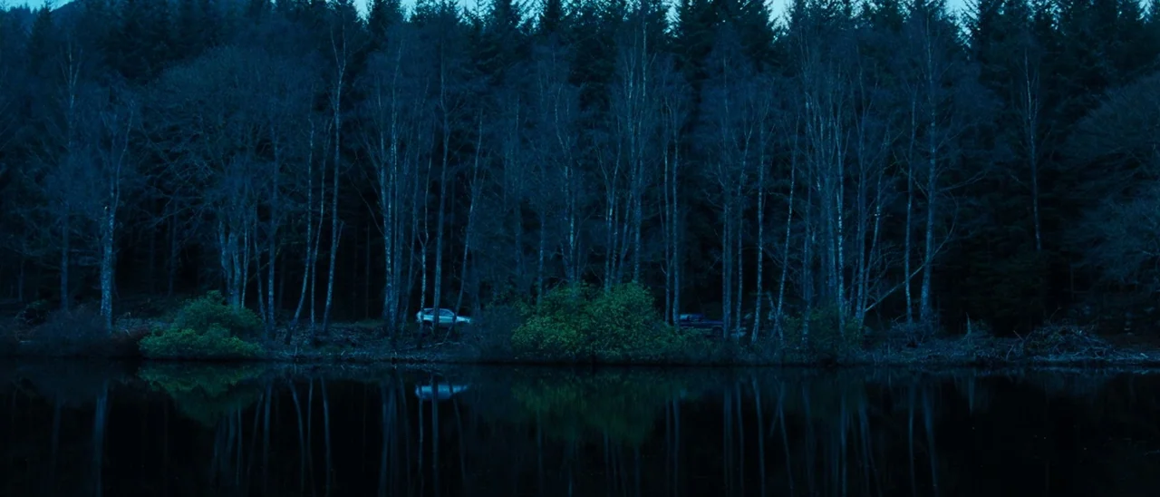 The Descent (2005) movie still: establishing — A dense wall of evergreen trees borders a calm, dark lake at night. Two cars are parked between the…