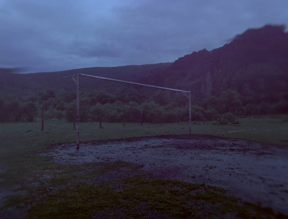Post Tenebras Lux (2012) movie still: establishing — A muddy soccer field sits at the base of dark, rolling hills under twilight. A weathered metal…