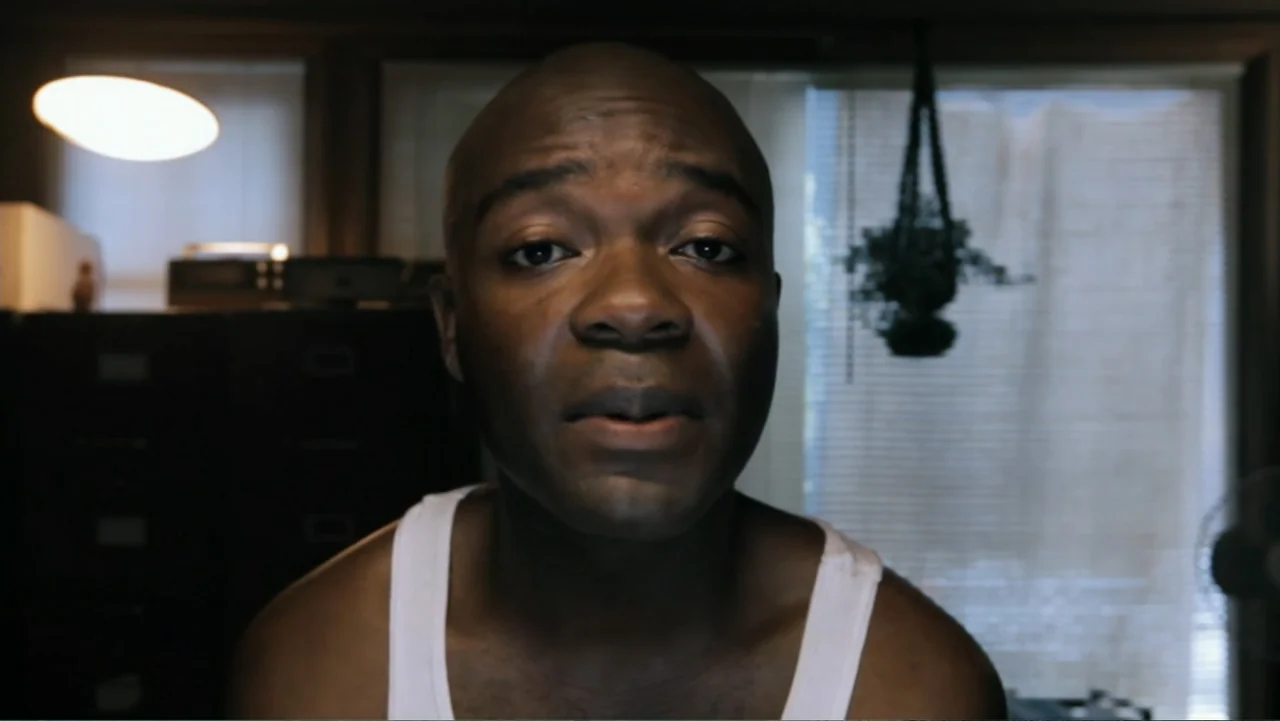 Nightingale (2014) movie still: closeup — An interior home office contains a dark filing cabinet and a window with blinds. A man in a white tank top…