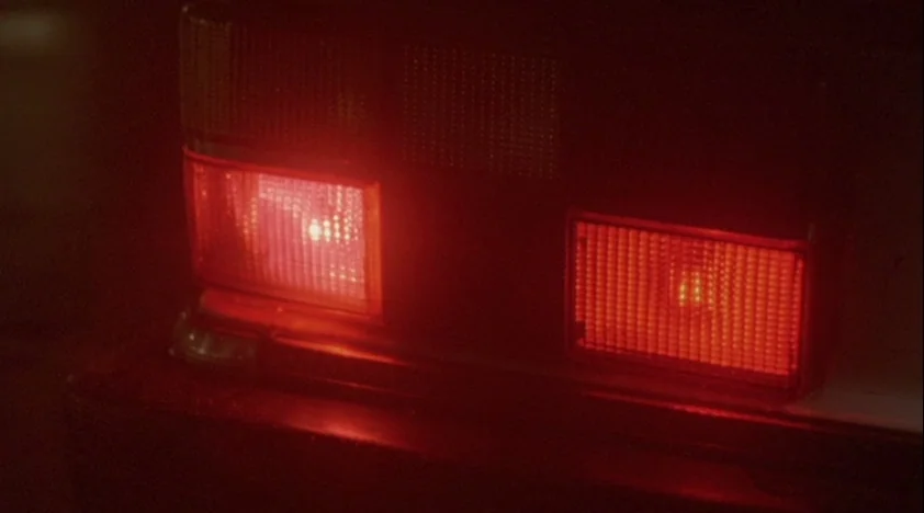 The House Of The Devil (2009) movie still: closeup — A close-up shot focuses on the illuminated red rectangular taillights of a car. The surrounding…