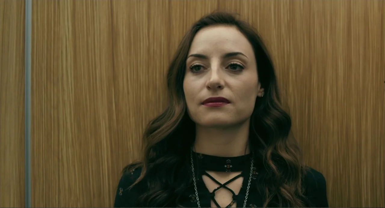 The Lift (2021) movie still: medium — A person stands against a smooth wood-paneled wall inside a modern lift. The subject faces the camera with a…