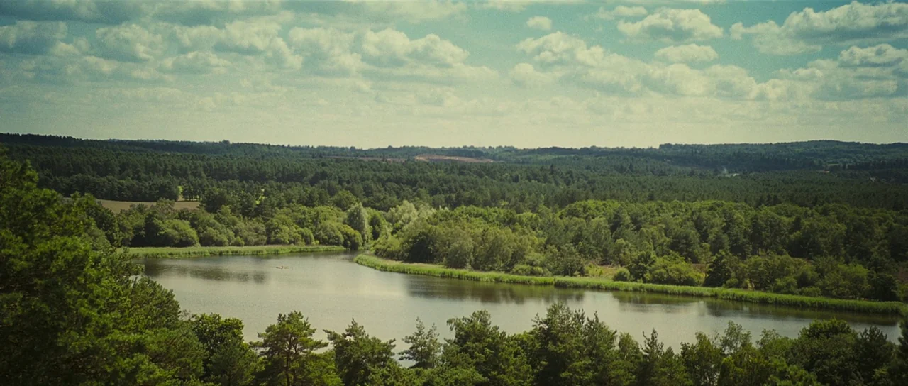 Eden Lake (2008) movie still: establishing — A wide, calm lake is surrounded by dense, rolling green woodlands and hills. The blue sky above is…