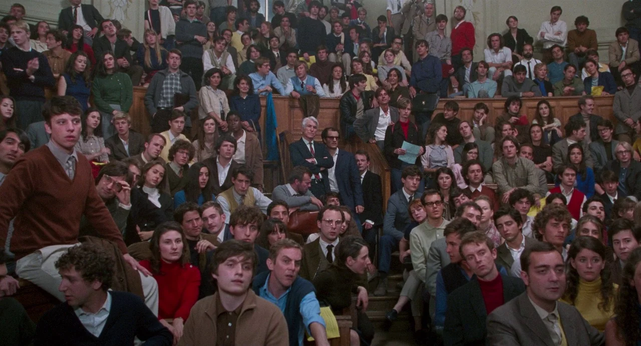 Godard Mon Amour (2015) movie still: wide — A tiered academic lecture hall filled with many students in 1970s clothing. People sit in wooden rows,…