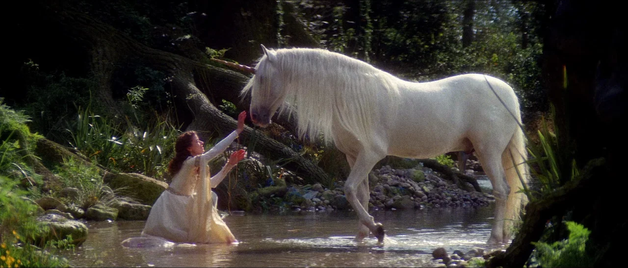 Legend (1985) movie still: wide — A shallow rocky stream flows through a lush, sun-dappled woodland with thick vegetation. A woman in a white gown…