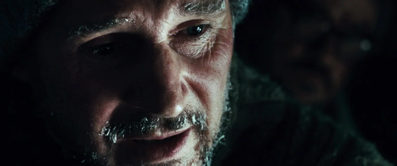 The Grey (2012) movie still: closeup — A man in a dark environment has ice gathered on his face. He gazes downward with a weathered expression while…