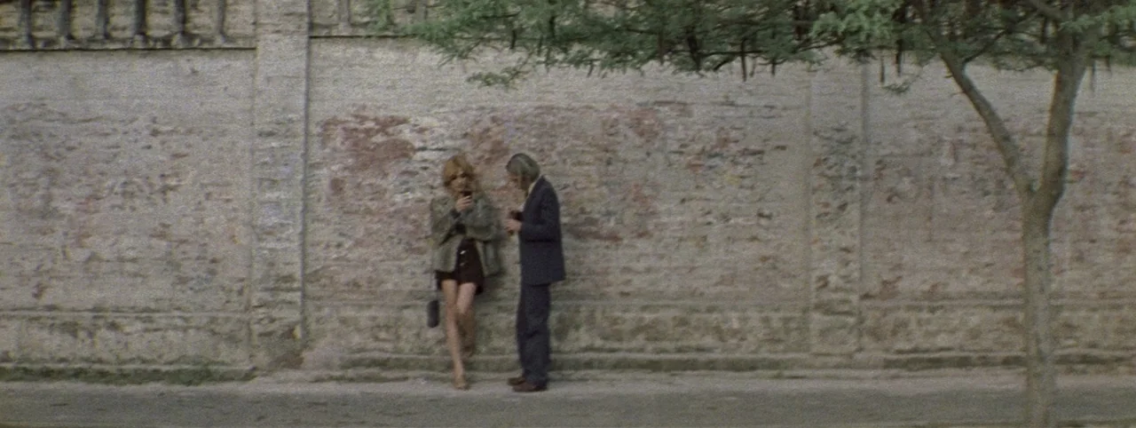 Post Mortem (2010) movie still: fullbody — A long weathered stone wall serves as a backdrop to a quiet outdoor sidewalk. Two people stand near the…
