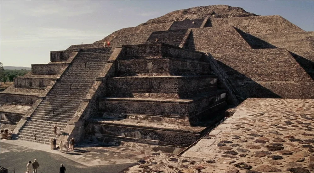 Frida (2002) movie still: establishing — A massive, multi-tiered ancient stone pyramid rises against a clear blue sky. Visitors wander at the base…