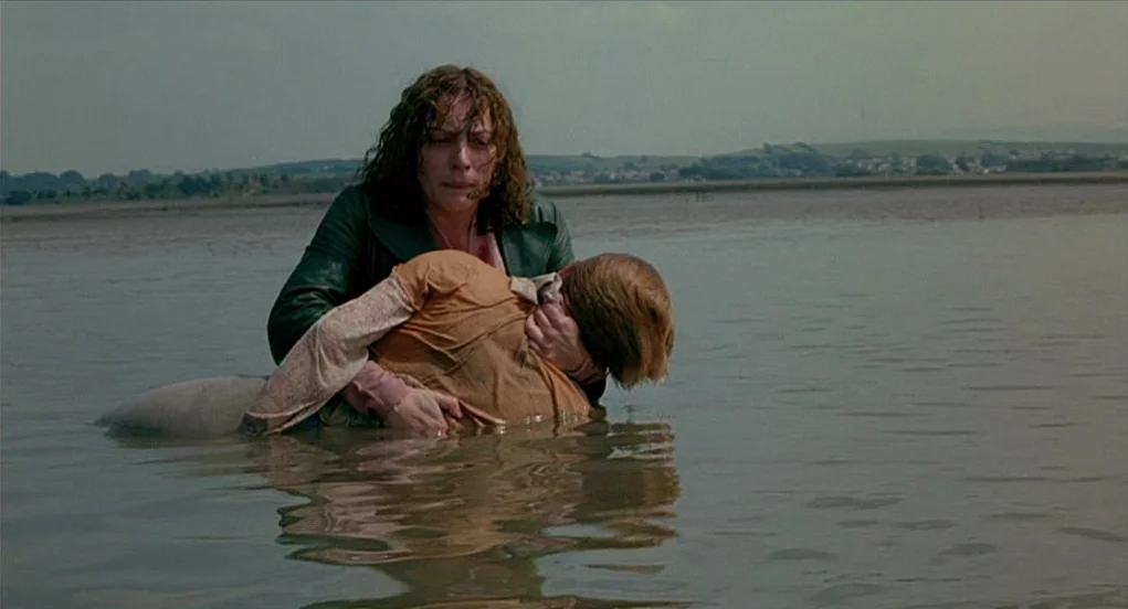 Butterfly Kiss (1995) movie still: medium — A woman stands waist-deep in shallow tidal water carrying an unconscious child. The horizon features…