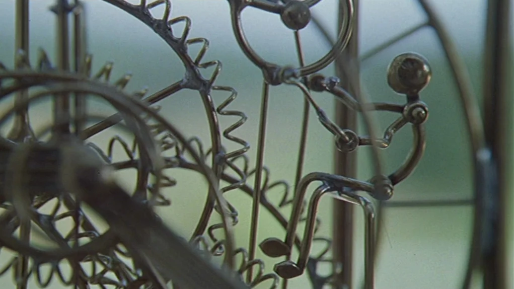 One Hundred and One Nights (1995) movie still: closeup — A close-up view shows a delicate wire figure integrated into a complex gear mechanism. The…