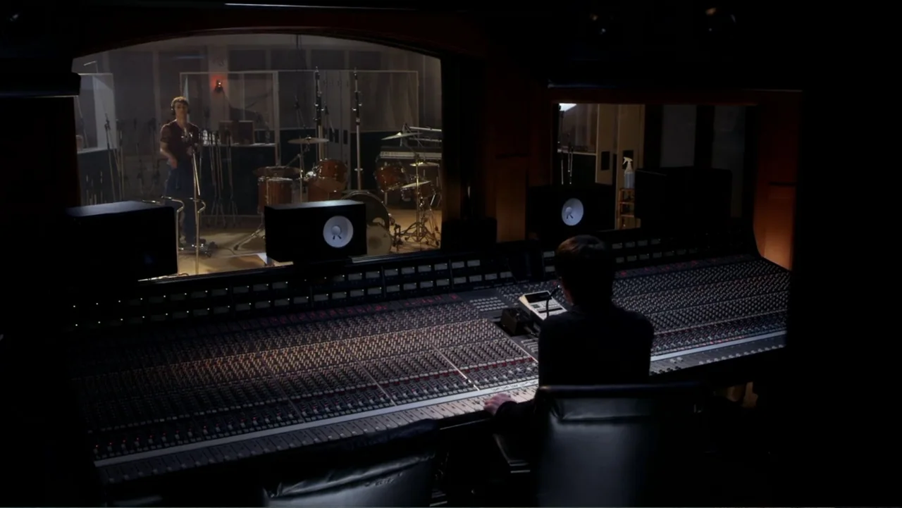 Rags (2012) movie still: wide — A professional music recording studio features a large mixing console in the foreground. A musician stands in a…