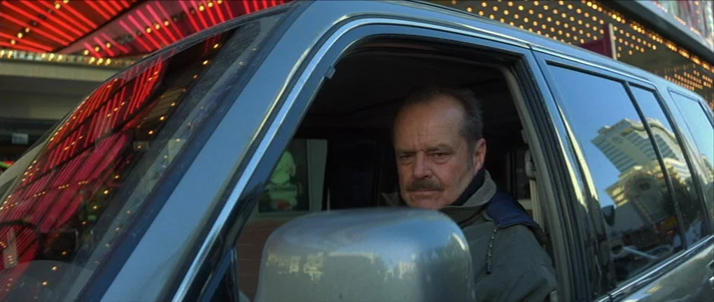 The Pledge (2001) movie still: medium — A man sits in the driver seat of a vehicle at night on a street. Bright neon lights from a nearby building…
