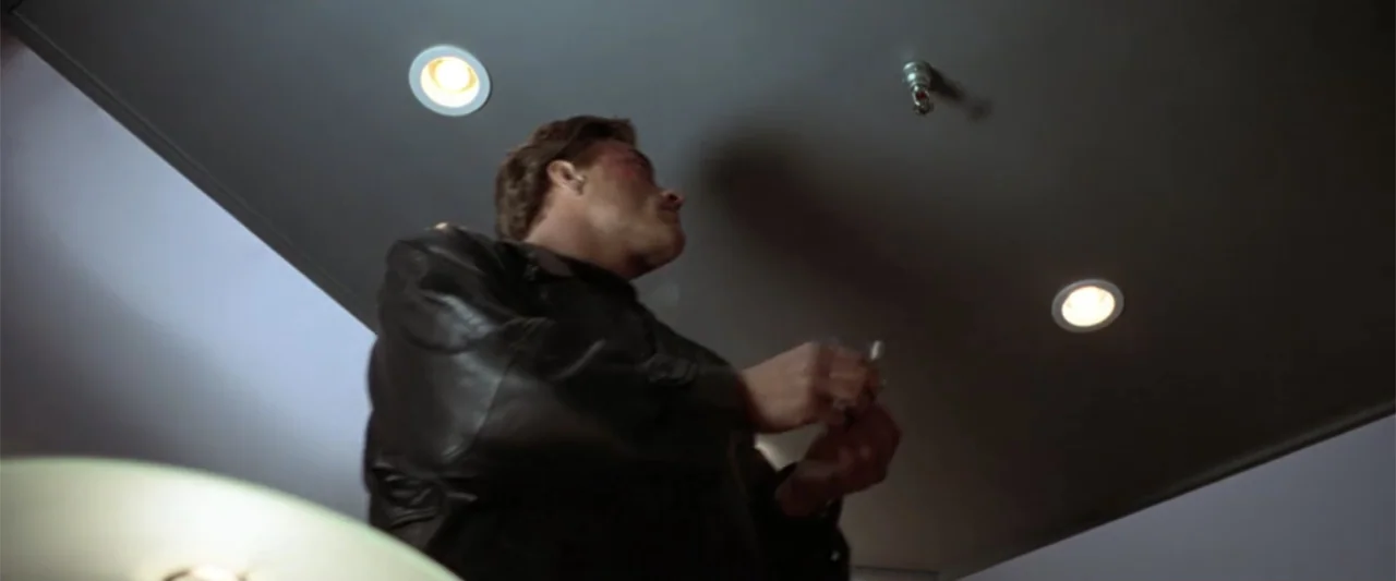 Maximum Risk (1996) movie still: medium — A man in a leather jacket stands inside a modern, gray-walled elevator. He looks up at the ceiling while…