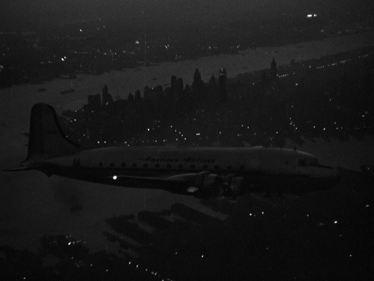 The Naked City (1948) movie still: wide — An American Airlines propliner flies over a city at night. The city skyline is visible in the background…