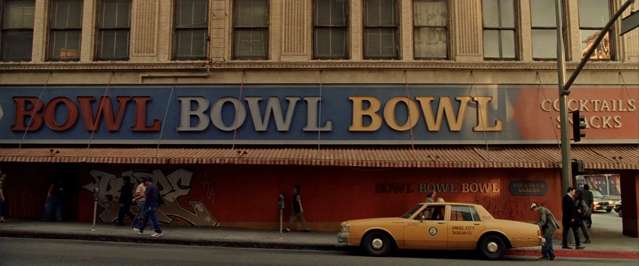 Constantine (2005) movie still: wide — A city street front features a large blue and orange sign reading Bowl Bowl Bowl. Pedestrians walk on the…