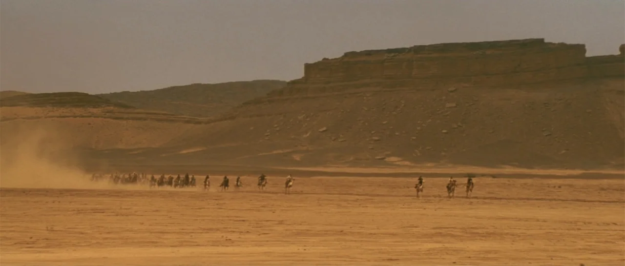 The Mummy (1999) movie still: wide — A vast, flat desert landscape stretches toward a large, eroded rocky mesa structure. A group of riders on…