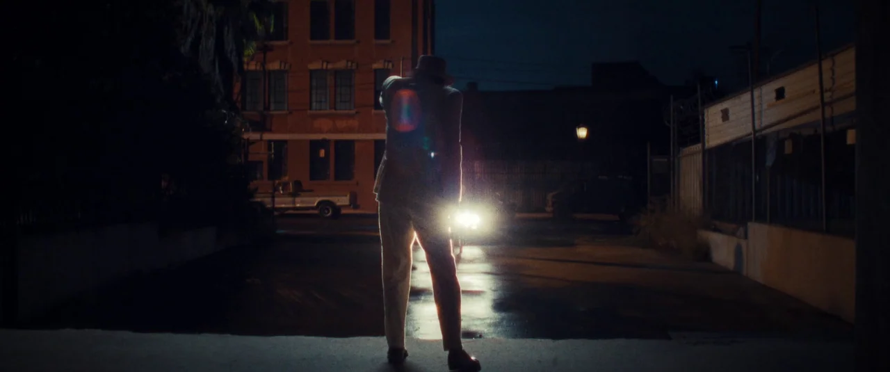 MaXXXine (2024) movie still: fullbody — An urban parking lot at night with a red brick building in the background. A man in a suit stands facing…