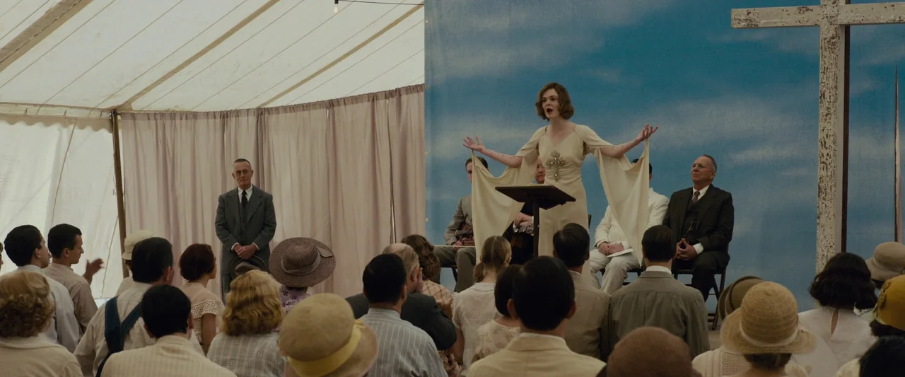 Live By Night (2016) movie still: wide — A woman stands behind a wooden podium inside a large fabric tent. She speaks to a seated audience in front…