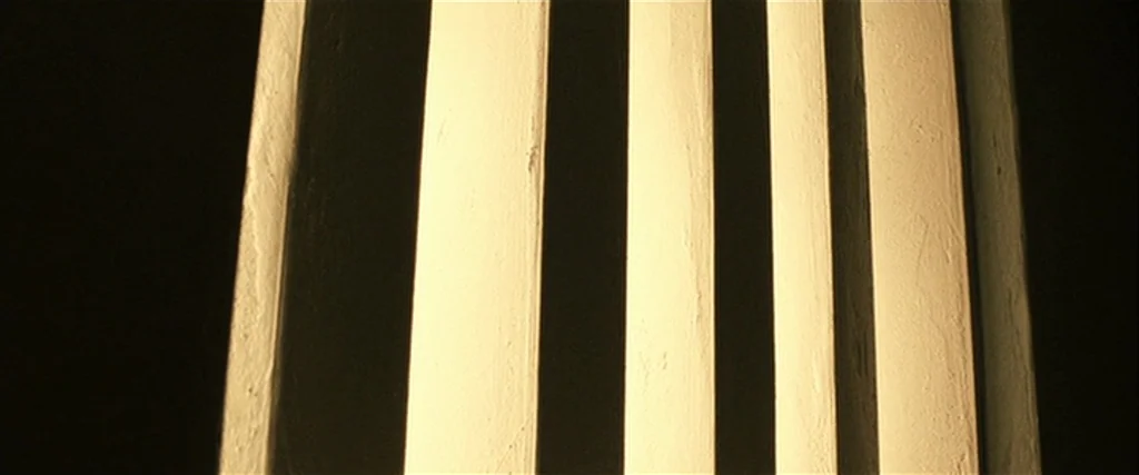 Hunger (2008) movie still: closeup — A close-up view shows the textured surface of several vertical architectural columns. Intense directional light…