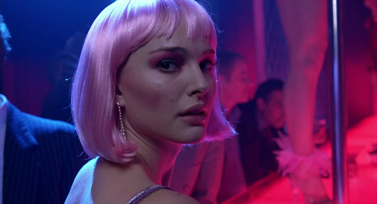 Closer (2004) movie still: closeup — A dark, dimly lit venue with bright neon lighting and background activity. A woman with a pink bob wig turns to…