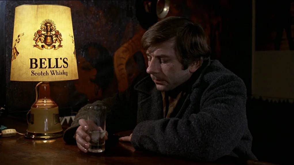 The Tenant (1976) movie still: medium — A dimly lit bar features a prominent branded lamp on a dark wood counter. The man sits slumped over his…