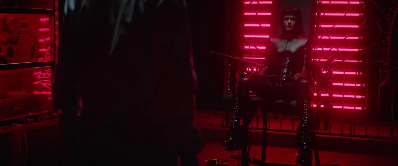 Dogs Don't Wear Pants (2019) movie still: medium — A dark, windowless room illuminated by vertical red neon bars mounted on a wall. A woman in latex…