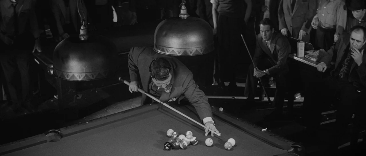 The Hustler (1961) movie still: medium — A dark, smoke-filled pool hall features large, low-hanging metal lamps over a green felt table. A man in a…