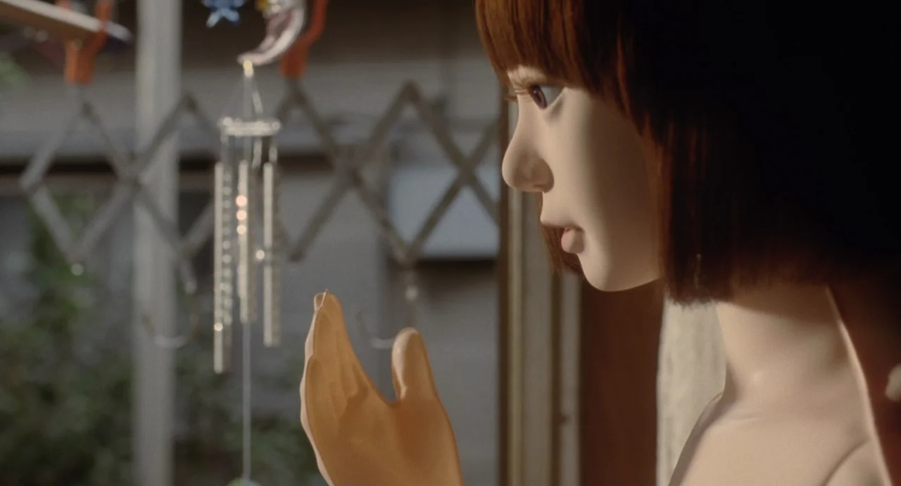 Air Doll (2009) movie still: closeup — An interior space with a window showing blurred outdoor foliage and a metal structure. A realistic humanoid…