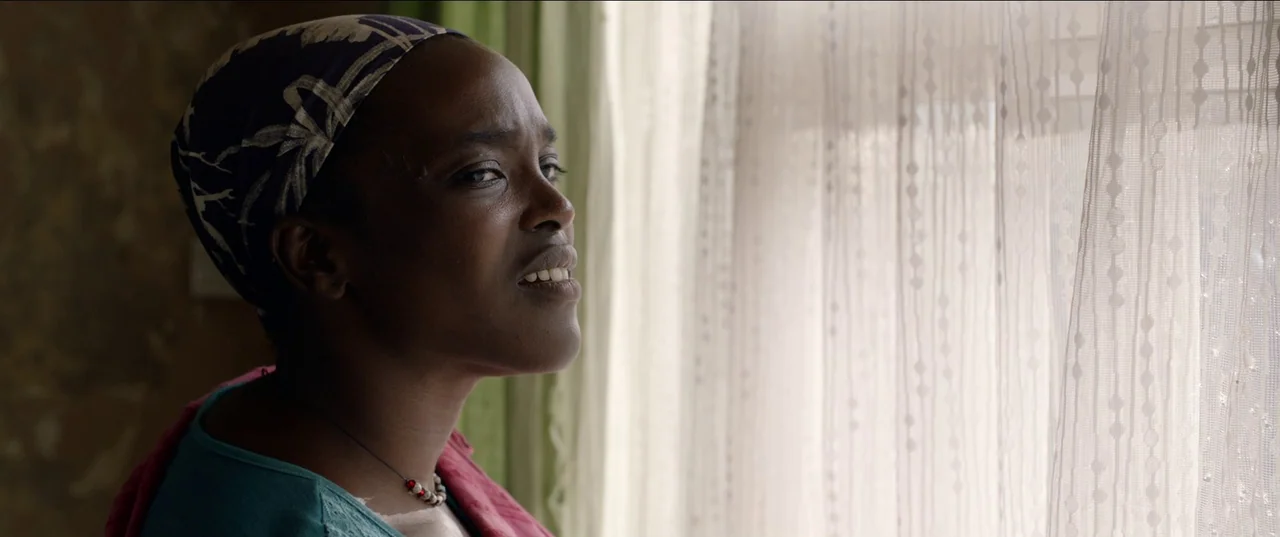 His House (2020) movie still: closeup — A woman stands indoors near a window partially covered by a thin sheer curtain. She looks outward with her…