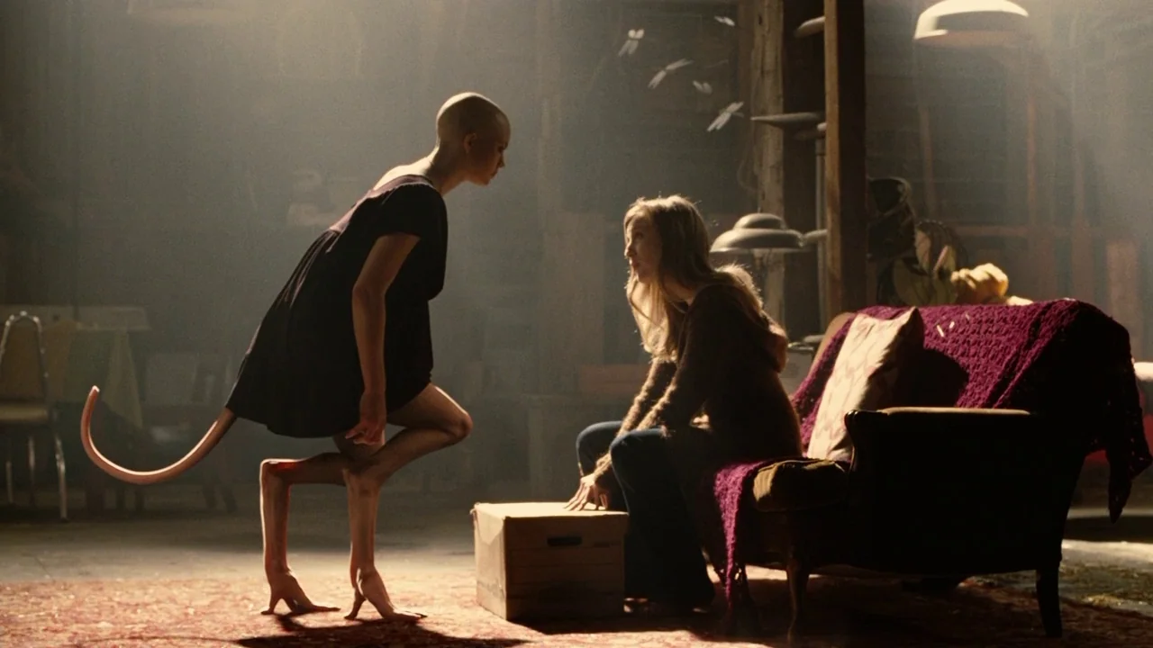 Splice (2009) movie still: medium — A dimly lit wooden attic contains a sofa covered with a purple throw. A humanoid creature with a tail faces a…