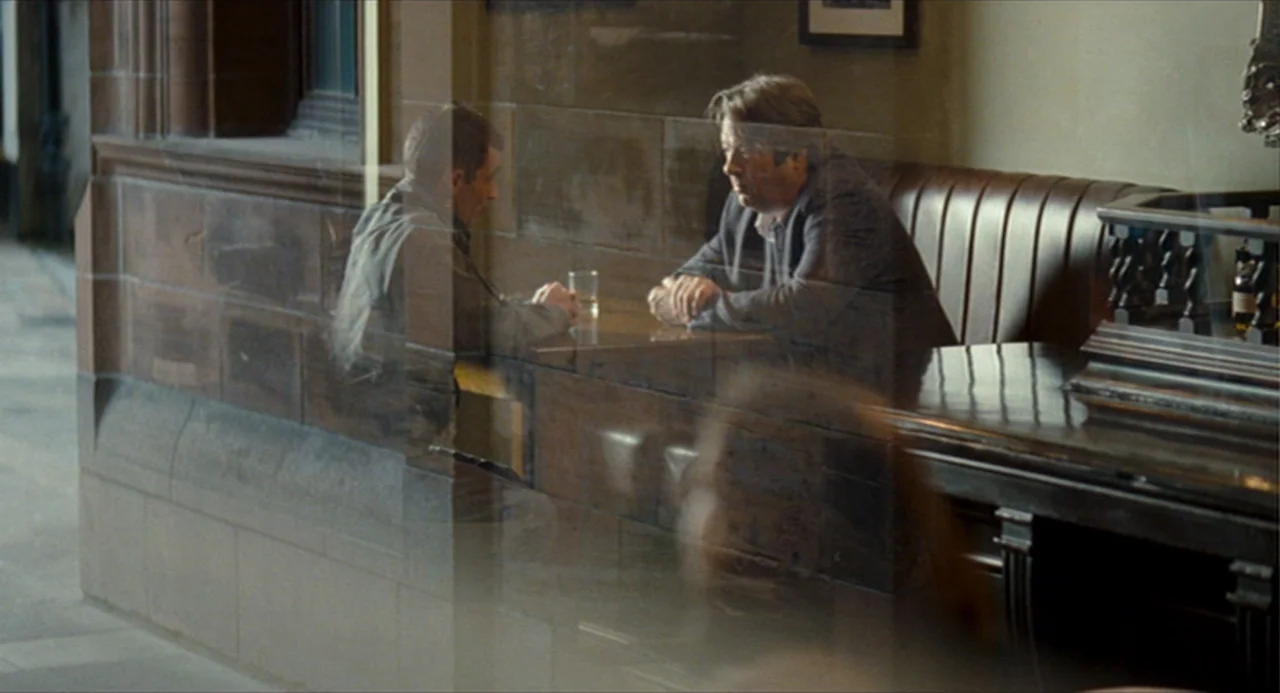 The Angel’s Share (2012) movie still: medium — A pub interior with dark wood paneling and leather booths seen through a window. Two men sit in a…