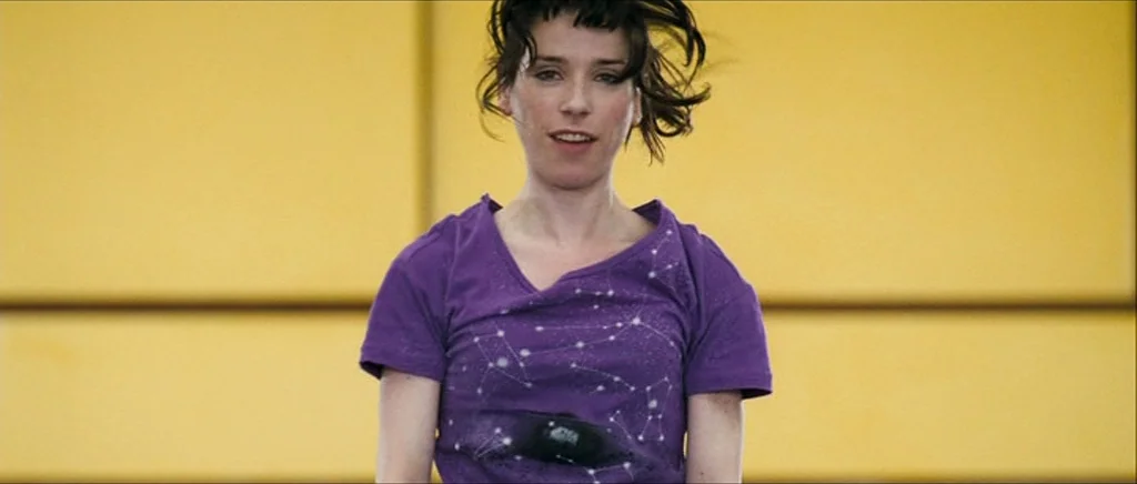 Happy-Go-Lucky (2008) movie still: medium — A woman stands in front of a solid yellow exterior wall with horizontal lines. She looks toward the…