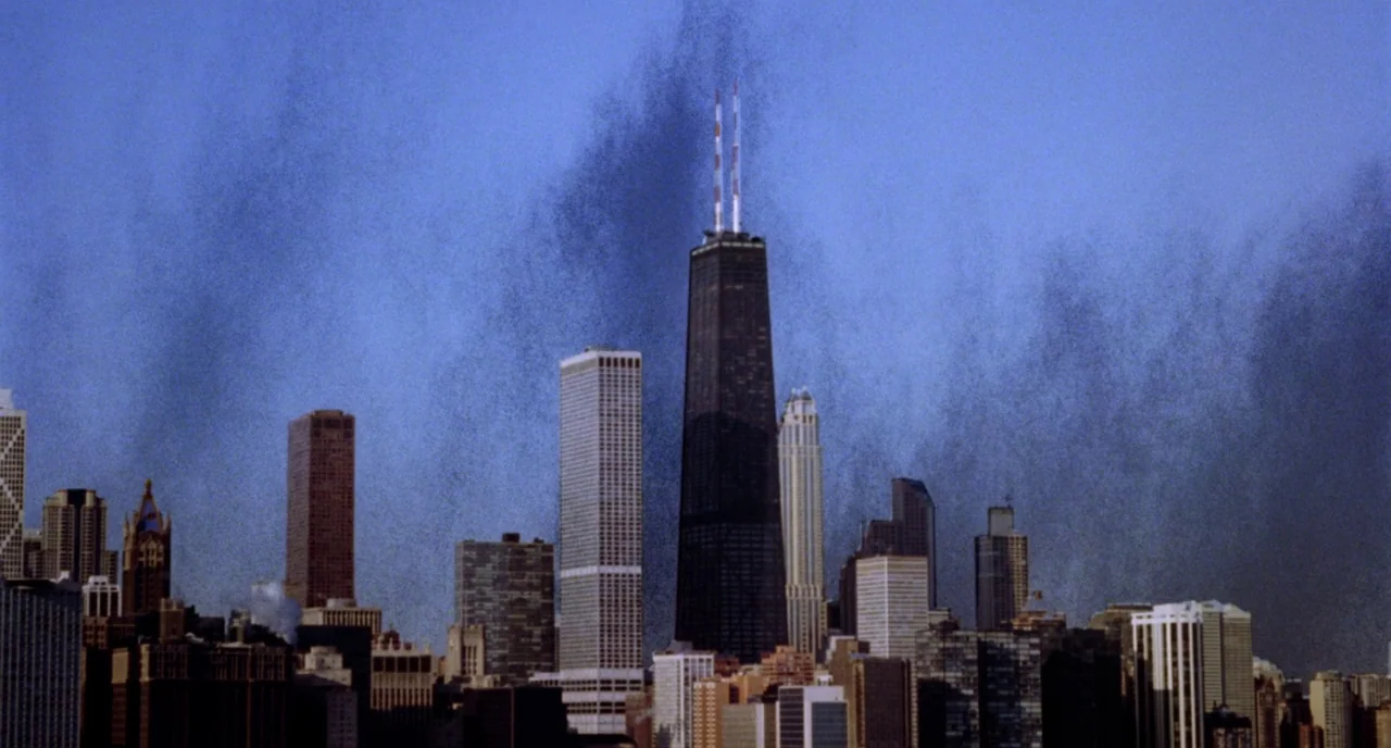 Candyman (1992) movie still: establishing — A dense urban skyline features multiple high-rise buildings against a clear blue sky. Thick plumes of…