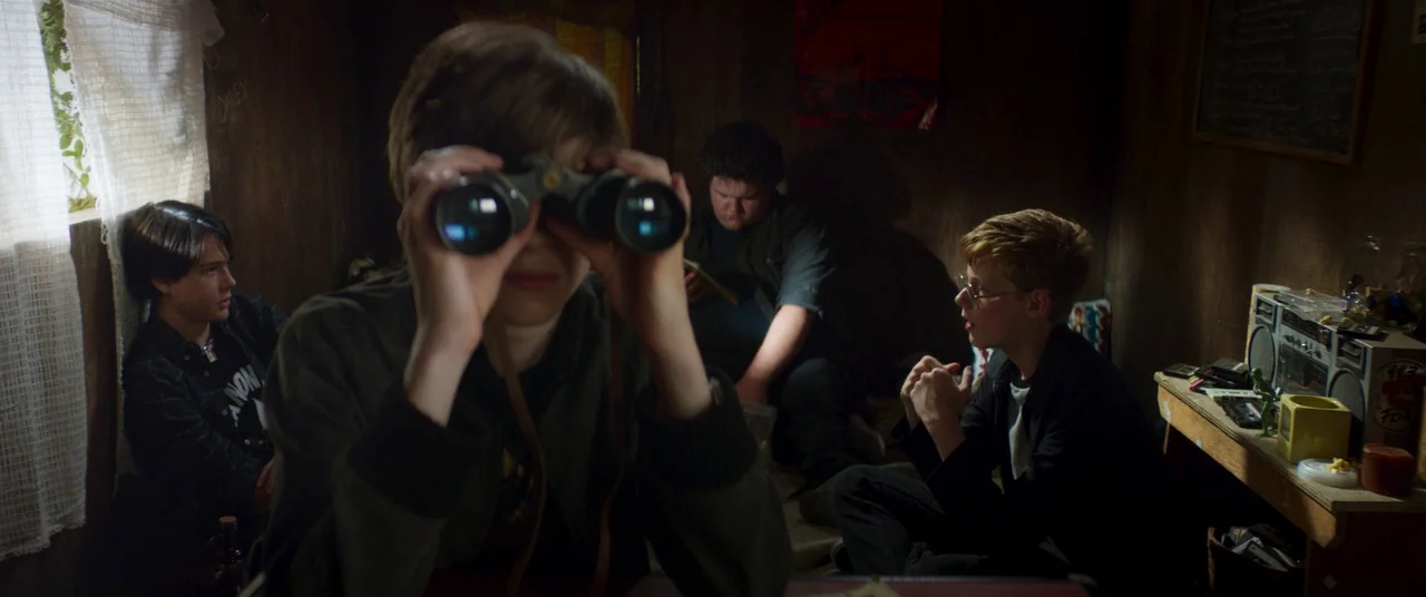 Summer of 84 (2018) movie still: medium — A dimly lit, wood-paneled room serves as a hideout for four young boys. The group sits in close proximity…