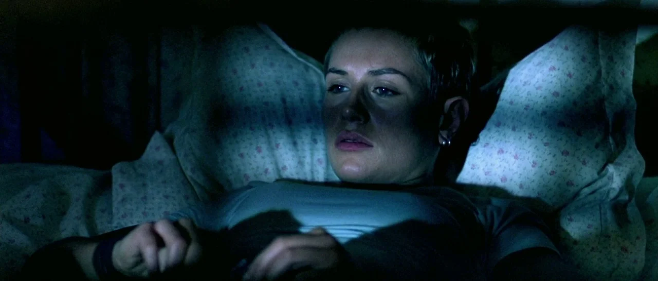 Haute Tension (2003) movie still: closeup — A dark bedroom with floral patterned pillows visible behind a person lying down. The person lies in bed…