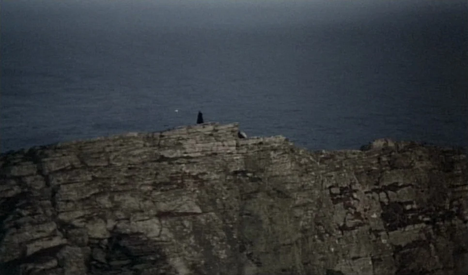 Heart of Glass (1976) movie still: wide — A rugged cliff face rises above dark, turbulent ocean waters under a grey sky. A small figure in dark…